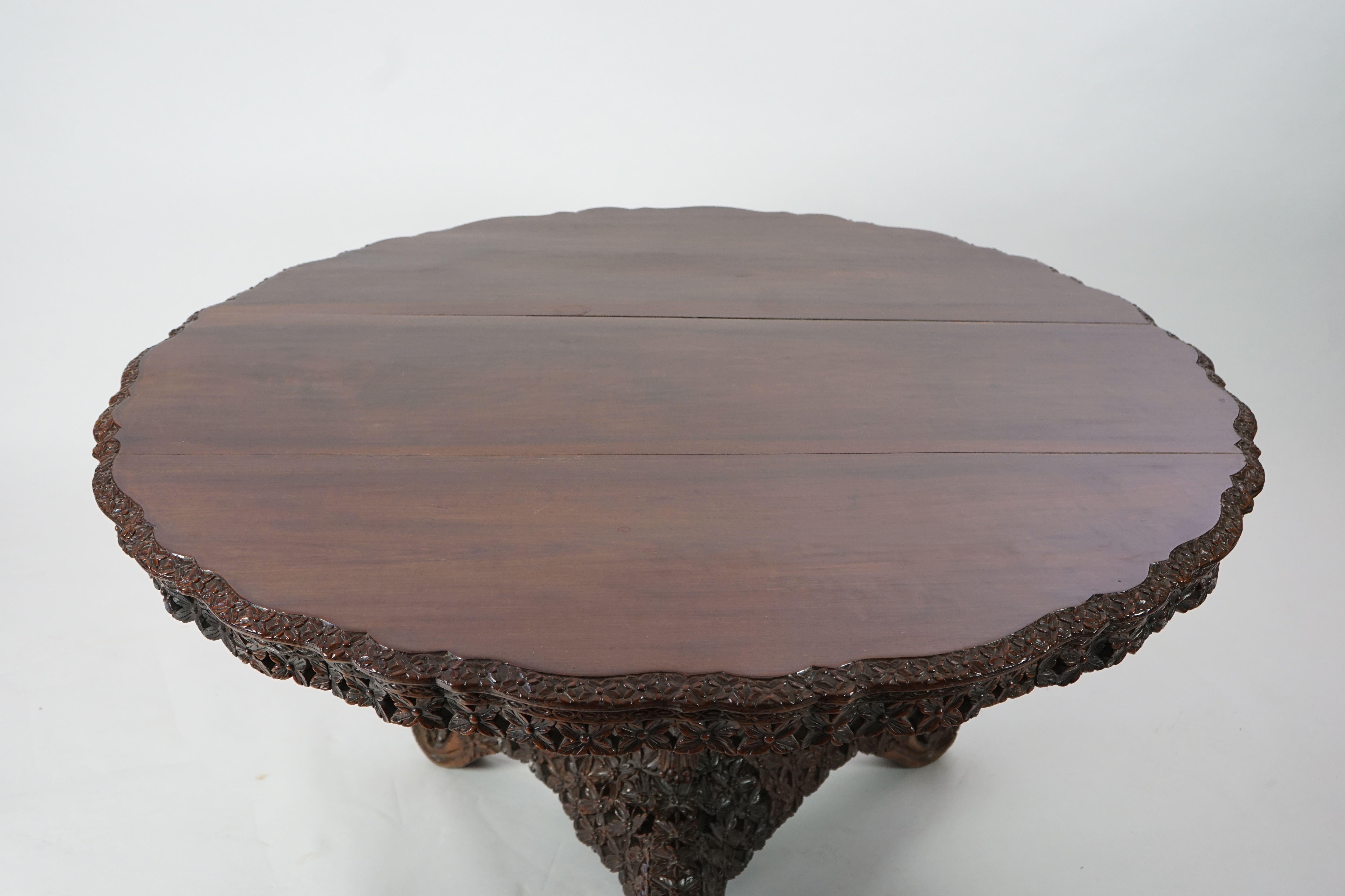A 19th century Anglo-Indian hardwood foliate carved tilt top centre table, with serpentine circular top, stylised floral carved rim and frieze, on heavily foliate and fruit carved pillar with pierced triangular platform